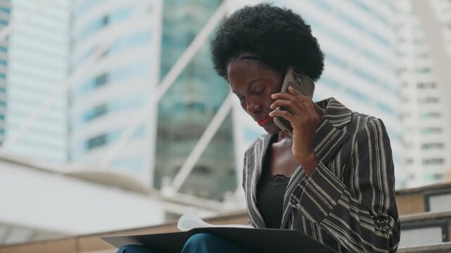 Professional woman talks on her phone. She sits outdoors, holding a tablet. Modern buildings loom behind her. Her focused expression suggests urgency