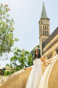 Caucasian Woman tourist wear white dress explores visit historic Christ Church Anglican Cathedral in Stone Town, Zanzibar. Features unique Gothic and Arabic coral stone in sunny day architecture