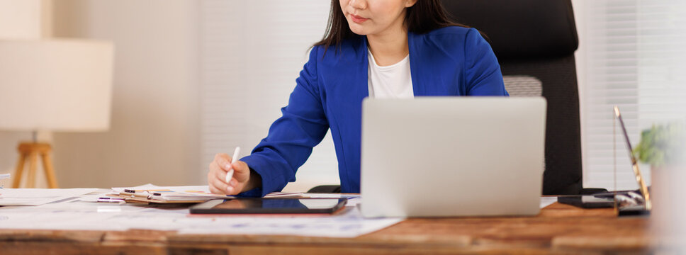 Young asian businesswoman in a blue blazer working on a project, using a tablet with a stylus pen and a laptop in a modern office, planning and analyzing data
