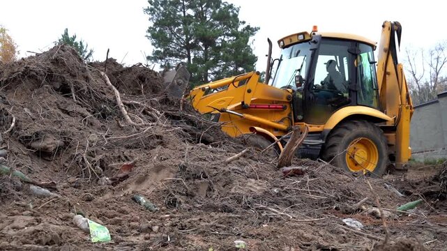 Yellow backhoe digs through soil and debris. Trees stand nearby, scattered leaves drift above. Heavy machinery moves earth, clearing messy piles. No workers visible, no human emotions shown