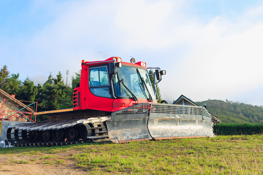 Red snow groomer parked on a grassy hill, waiting for winter, showcasing ski resort maintenance equipment against a clear sky and distant buildings
