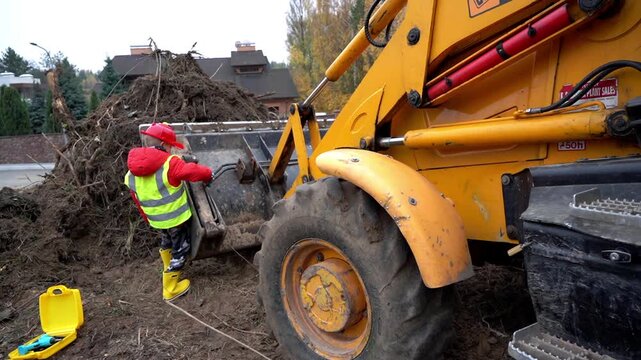Worker in bright gear directs heavy machinery clearing debris. Yellow loader moves large piles of soil and branches. Scene shows urban cleanup afterstorm damage