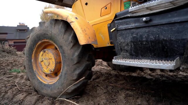 Yellow tractor tire crunches through muddy soil. Large tread marks show heavy machinery work. Background shows autumn trees and a rustic house. Ground is freshly turned, dirt scattered
