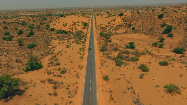 Single vehicle travels along a straight paved highway through dry Thar Desert dunes and sparse shrubs, viewed from a high moving drone.