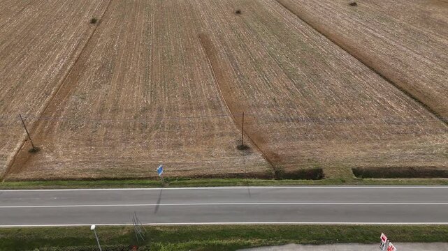 Harvested cereal field stretches beside a rural road in Italy, with dense stubble rows, field tracks and utility poles defining the agricultural landscape, aerial slide descent