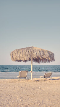 Straw beach umbrella and lounge chairs on a sandy beach, color toning applied.