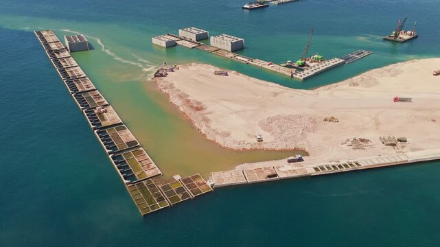 Industrial machinery and barges working on a large-scale coastal engineering project, building a breakwater and reclaiming land from the sea with caissons for a new port development