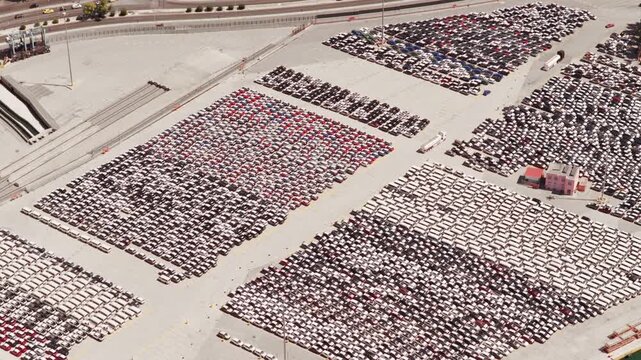 High-angle view of a vast parking lot in an industrial port, packed with thousands of new cars sorted by color and ready for shipping, distribution, and export to the global market