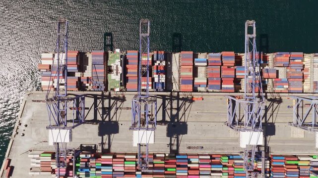 High angle drone footage showing a huge cargo ship arriving at a modern seaport and mooring at the pier for loading and unloading operations with gantry cranes moving over containers