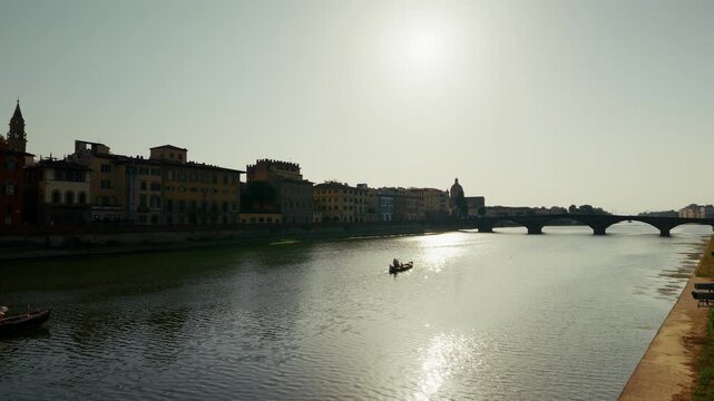 A single sculler crosses the sun's reflection on the Arno with moored boats waiting along the Lungarno bank in Florence, Tuscany. Static backlit wide shot from a bridge.