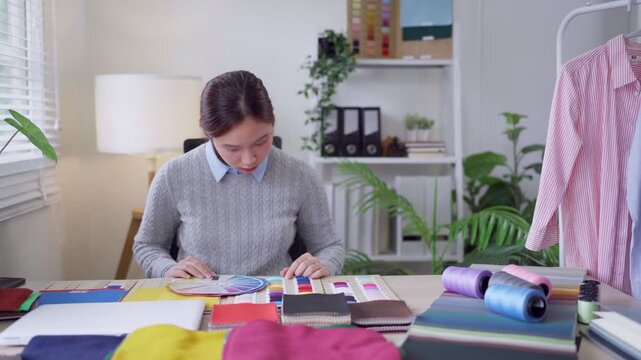 Young Asian fashion designer examining color swatches and fabric samples at cluttered studio desk