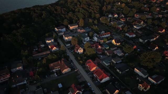 Red rooftops glow beneath golden hour light in Usedom coastal town, dense pine forest borders quiet residential streets, Baltic Sea stretches beyond distant tree line. Aerial drone flyover shot