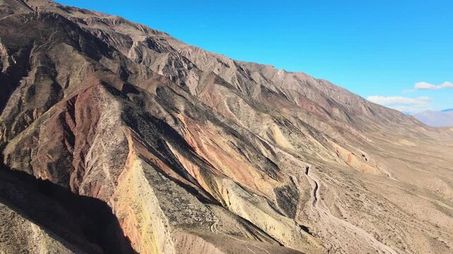 Multicolored rock ridges of Paleta del Pintor descend toward a dry valley under clear blue sky, seen from a high drone flyover.