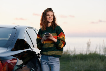 Obraz premium A smiling young woman holding a smartphone stands beside her car at sunset. Concept for modern travel, road trip lifestyle, and mobile communication during a journey.