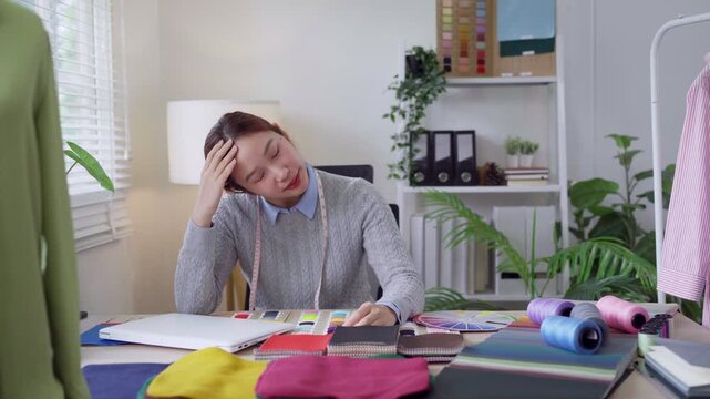 Stressed young Asian fashion designer rubbing temples overwhelmed by fabric swatches at studio desk