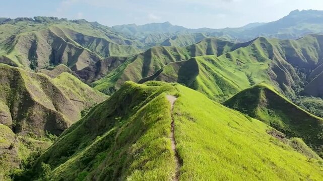 Aerial view of a hiker walking on a narrow ridge path across lush green rolling hills and mountain ranges in a tropical landscape