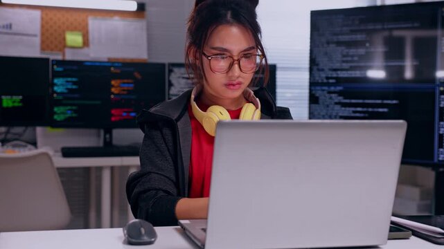 Young Asian woman programmer using laptop modern software development workspace with computer monitors, colorful code screens, focused expression, technology programming and digital development work