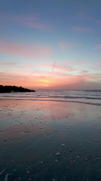 Vertical video of a serene beach at sunset with calm waves and beautiful pink sky reflected on the wet sand.