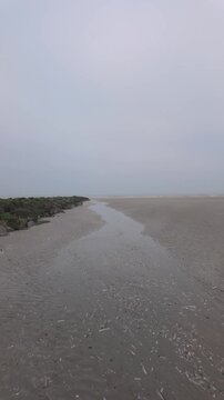Moody vertical footage of walking along a desolate, foggy beach with a stream flowing towards the sea.