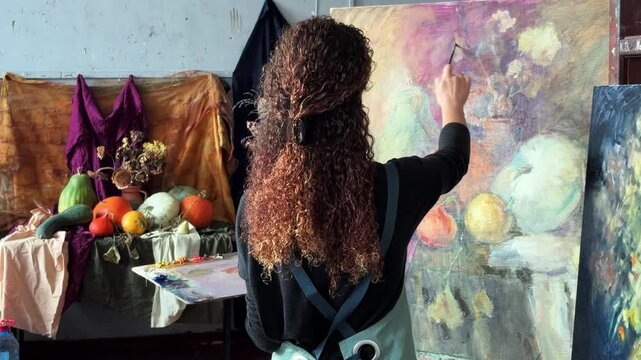 Teenage female artist in college painting class, art studio, art workshop. She paints still life on canvas with oil paints, holds brush, palette.