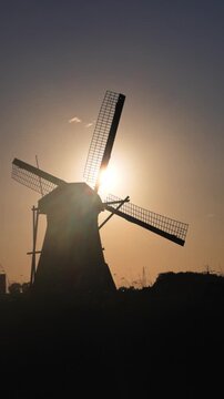 Silhouette of a traditional Dutch windmill with rotating blades against a beautiful golden sunset sky. Vertical footage.