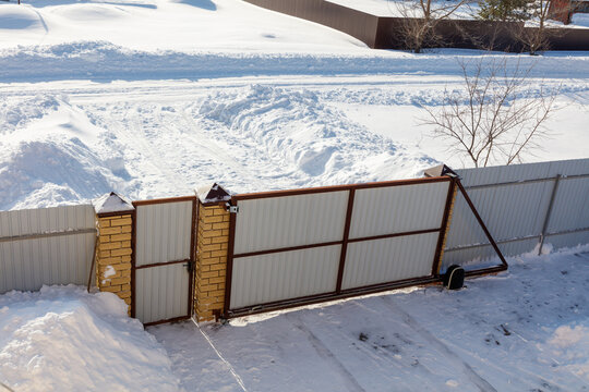 Snow covers a winter yard with a leaning gate. Metal gate leans on a snow-lined fence.