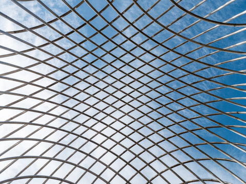 Abstract view of a curved metal lattice structure against blue sky, creating a geometric grid of intersecting lines for architecture, engineering and structural design concepts.