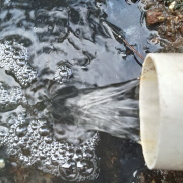 Water flows rapidly out of a white PVC pipe into a foaming, bubbling puddle on the wet ground or rocks.w