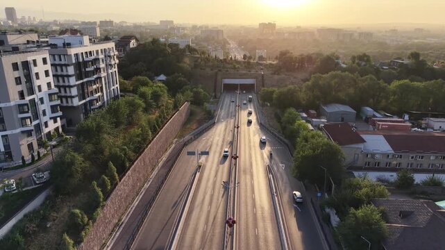 Drone view flying over busy freeway into bright sunset sun with urban park and cityscape