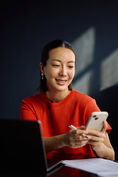 Happy Korean woman using cell phone app while working at home.