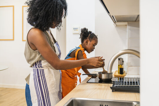 African american mother and daughter wearing aprons, stirring a pot on the stovetop, learning to cook in a modern home kitchen