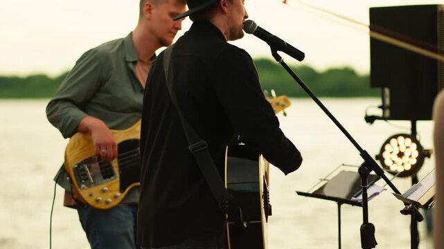 Outdoor riverside live music performance with guitarist singing into microphone and bassist accompanying on stage under open sky during relaxed evening concert