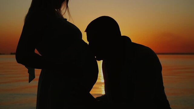 Expectant father kisses his partners pregnant belly and then listens to the baby, creating a tender silhouette of parenthood, pregnancy and family bonding at sunset.
