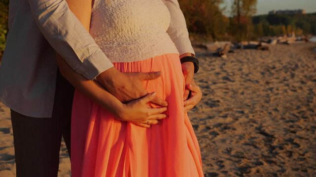 Close-up of expecting parents gently holding a pregnant belly together on the beach at sunset, expressing love, support, family bonding and anticipation of their baby.