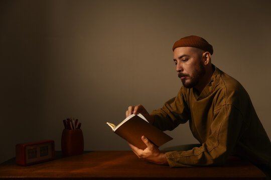 Focused man reading a book while sitting at a wooden desk with a vintage clock, wearing a beanie and enjoying quiet study, learning, or literature in a cozy, warm, and atmospheric room setting.