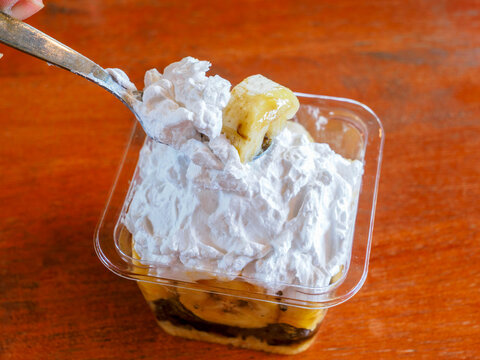 Close-up of spoon scooping banoffee pie cup topped with fluffy whipped cream and banana slices in clear plastic container.