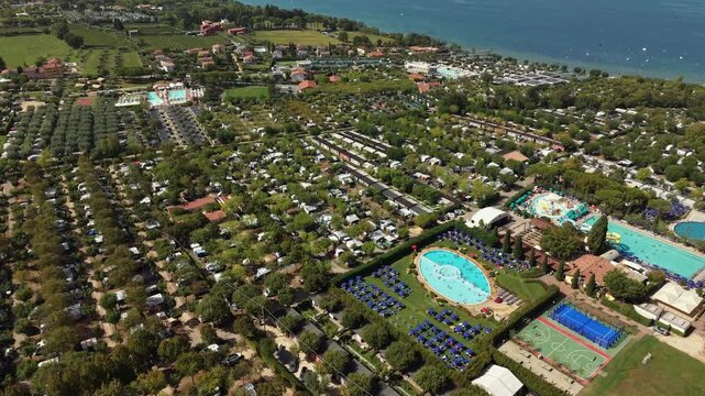 Circular swimming pool anchors resort courtyard lined with sun loungers, water park and tennis courts border dense rows of camper vans near shoreline of Lake Garda, Italy. Aerial drone flyover shot