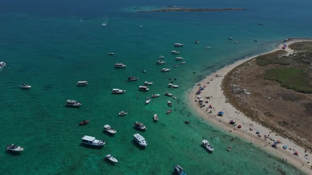 An aerial drone shot over the clear turquoise sea, sandy beach, and rocky coastline of Rabbit Island in Tripoli, Lebanon, featuring anchored boats and swimmers.