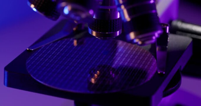 Microscope inspecting circular silicon wafer with microchip on laboratory stage with purple and blue lighting.