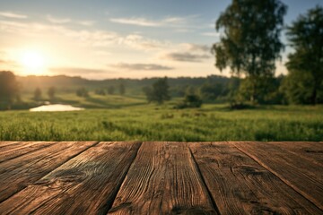 Obraz premium A rustic wooden table overlooks a serene countryside landscape with green fields, trees, and a glowing sunrise or sunset in the background.