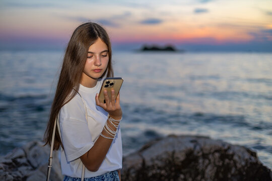 Young Woman Using Smartphone by Sea at Sunset. Young woman looks at a smartphone while standing beside a rocky seashore at sunset with calm water and a colorful sky.