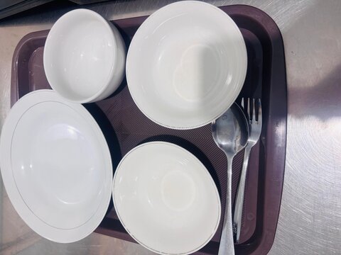Top-down view of an empty white ceramic tableware set including bowls, a plate, spoon, and fork arranged on a brown plastic tray.
