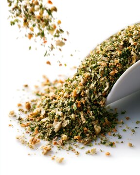 A close-up of dried mixed herbs and spices spilling from a white spoon onto a white surface.
