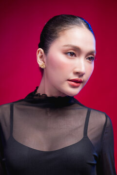 A poised beauty portrait with a direct, symmetrical gaze, sleek center-parted hair and sheer black attire against a rich red backdrop, exuding calm confidence.