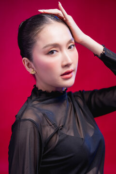 An artistic portrait with an arm curved gracefully overhead, gaze lifted contemplatively, elongated neckline and sheer black fabric creating a flowing silhouette against deep red.