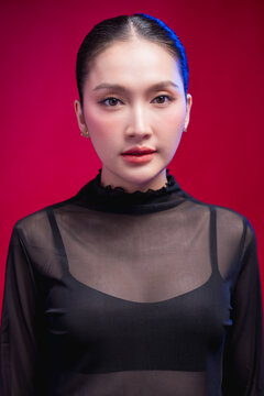 A poised beauty portrait with a direct, symmetrical gaze, sleek center-parted hair and sheer black attire against a rich red backdrop, exuding calm confidence.