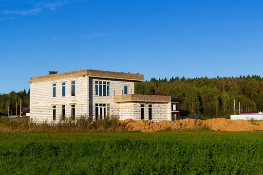 Unfinished two-story residential house made of white silicate blocks under construction in a picturesque rural field with a forest in the background under a bright clear blue sky
