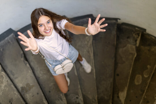 Smiling Young Woman Reaching Up on Spiral Staircase. Smiling young woman looks up and reaches both hands toward the camera while standing on a dark spiral staircase.