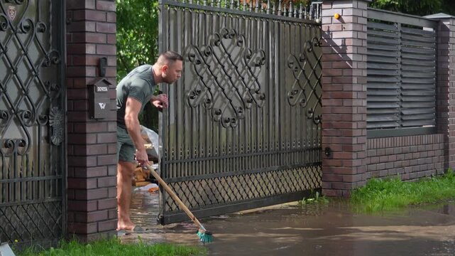 Man uses a heavy squeegee broom to forcefully push muddy floodwater out of his suburban private house driveway.