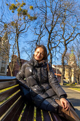 Smiling Woman Sitting on Park Bench in Winter Sunlight. Smiling woman in a shiny black puffer jacket sits on a wooden park bench in warm sunlight beneath leafless trees in an urban park. © Iryna